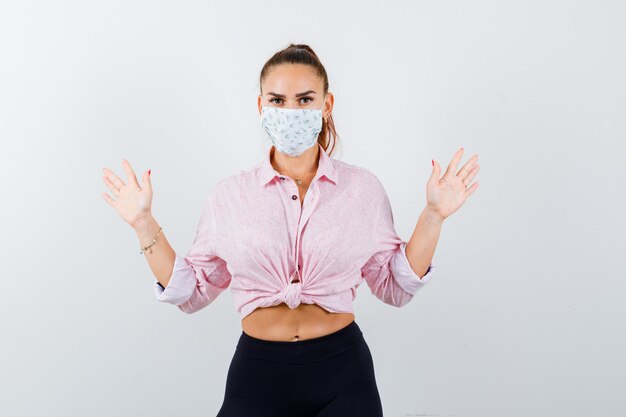 Jeune fille levant les paumes en geste de capitulation en chemisier rose, pantalon noir, masque et air effrayé, vue de face.