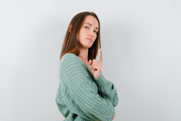 Jeune fille en maille debout les bras croisés, regardant par-dessus l'épaule, levant l'index en geste eureka et regardant sensible, vue de face.