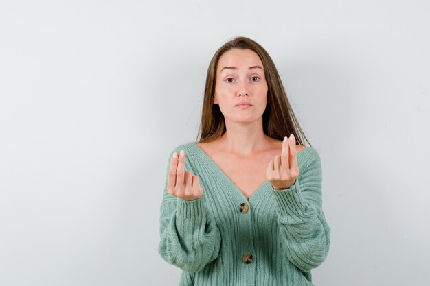 Jeune fille en maille montrant le geste de l'argent et à la vue sérieuse, de face.