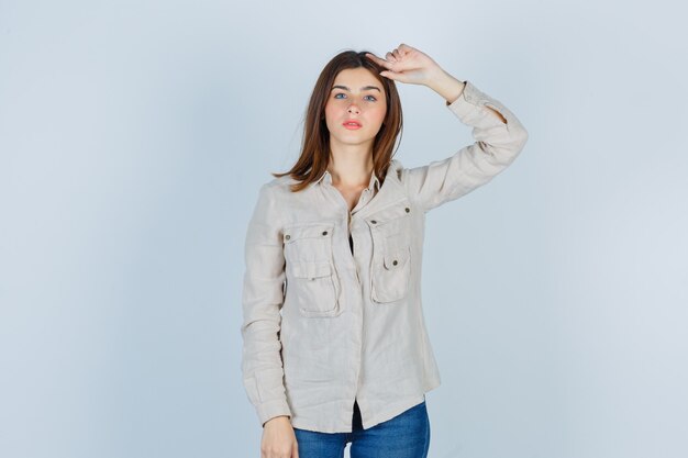 Jeune fille avec la main sur la tête en chemise beige, jeans et l'air mignon. vue de face.