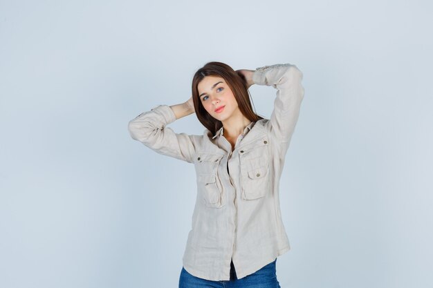 Jeune fille avec les mains derrière la tête tout en posant en chemise beige, jeans et charmante, vue de face.