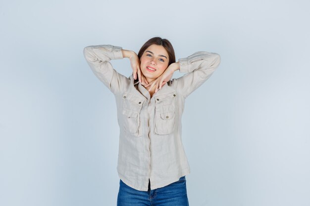 Jeune fille avec les mains sous le menton en chemise beige, jeans et l'air mignon. vue de face.