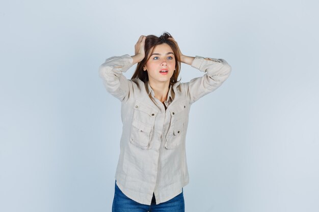 Jeune fille avec les mains sur la tête en chemise beige, jeans et l'air surpris. vue de face.