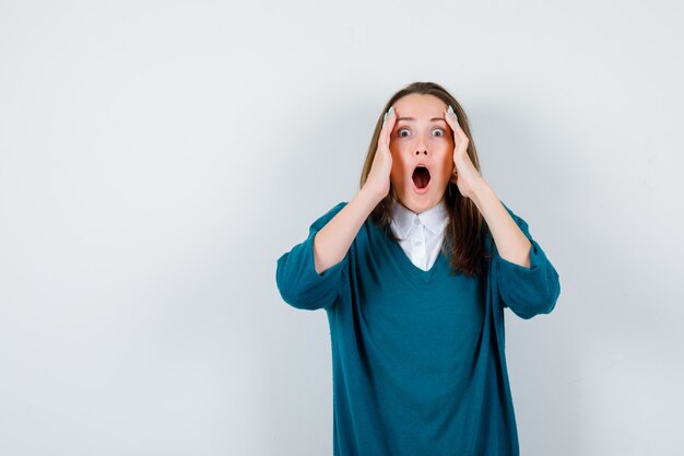 Jeune fille avec les mains sur le visage, ouvrant la bouche dans un pull à col en V, une chemise et l'air choqué, vue de face.