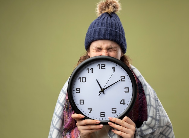 Photo gratuite jeune fille malade en colère portant robe blanche et chapeau d'hiver avec écharpe visage couvert avec horloge murale enveloppée de plaid isolé sur vert olive