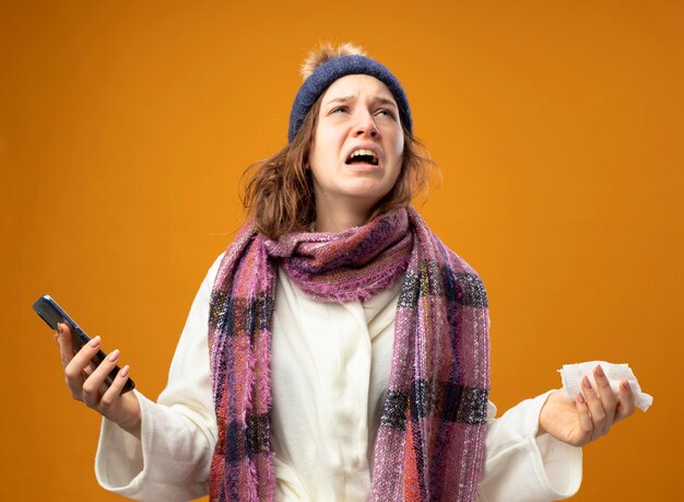 Photo gratuite jeune fille malade en colère portant une robe blanche et un chapeau d'hiver avec foulard tenant le téléphone et la serviette étalant les mains isolés sur le mur orange