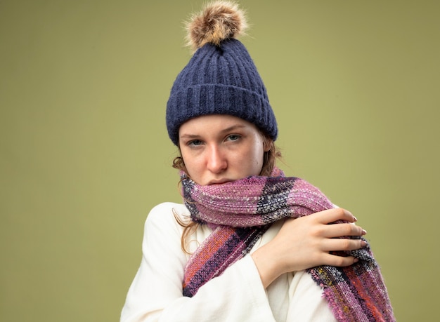 Jeune fille malade regardant droit devant portant une robe blanche et un chapeau d'hiver avec écharpe mettant la main sur l'épaule isolé sur vert olive