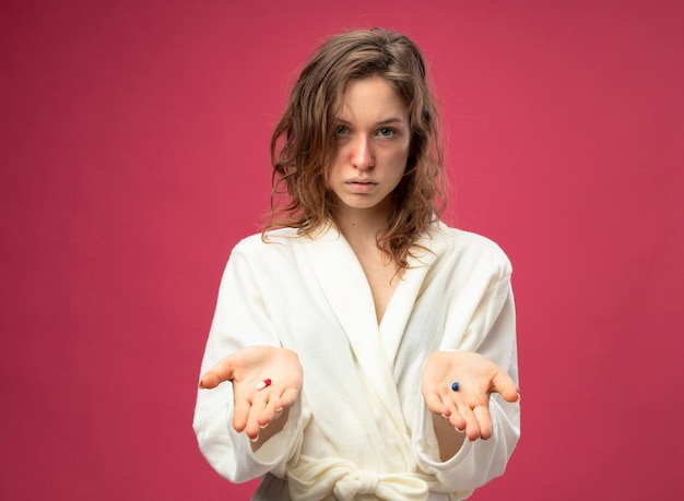 Jeune fille malade regardant droit devant portant une robe blanche tenant des pilules isolé sur rose