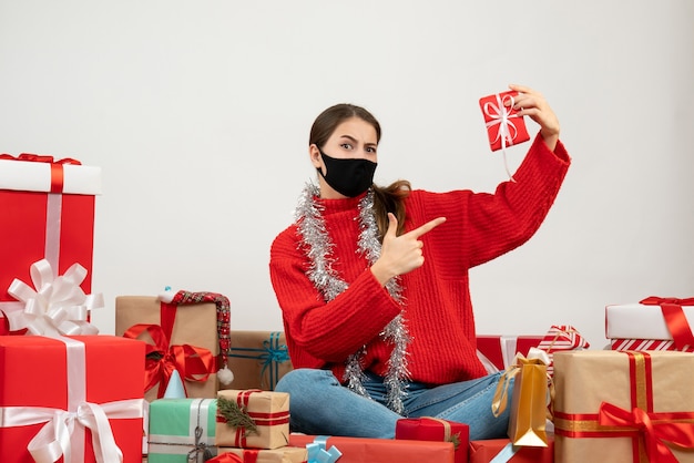 Jeune fille avec un masque noir pointant sur cadeau assis autour de cadeaux sur blanc