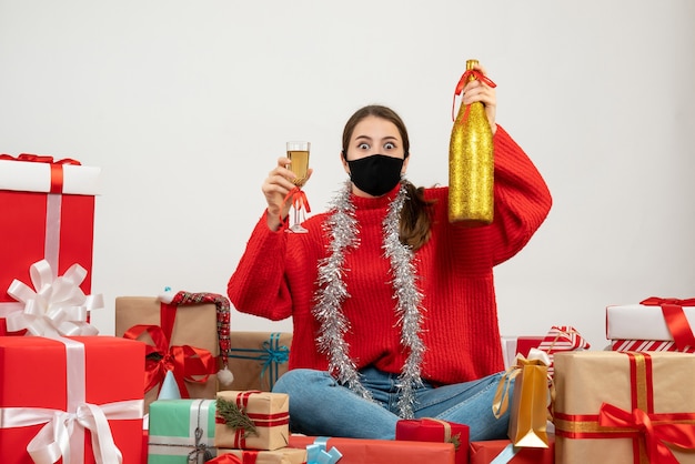 Jeune fille avec masque noir tenant une bouteille de champagne et un verre assis autour de cadeaux sur blanc