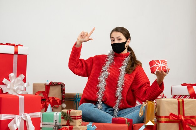 Jeune fille avec masque noir tenant cadeau faisant signe de pistolet doigt assis autour de cadeaux sur blanc