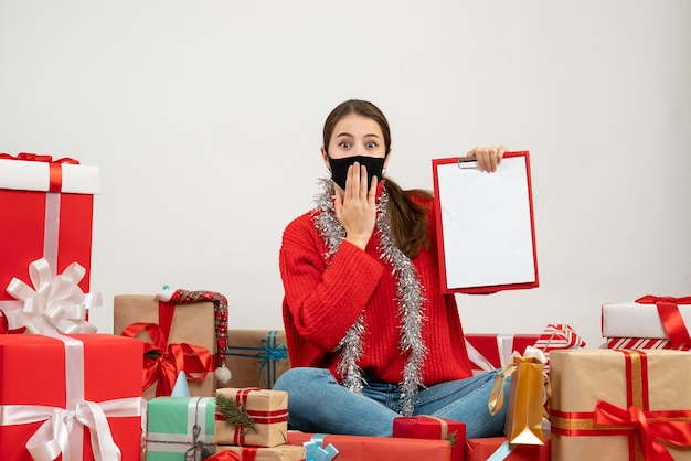 Jeune fille avec masque noir tenant des documents assis autour de cadeaux sur blanc