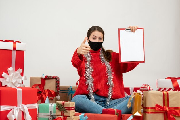 Jeune fille avec masque noir tenant des documents faisant le pouce vers le haut signe assis autour de cadeaux sur blanc