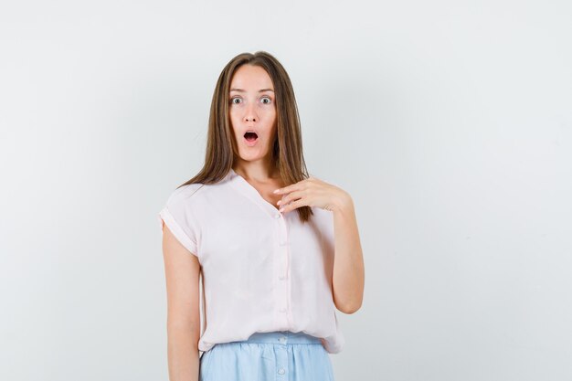 Jeune fille me faisant signe? en t-shirt, jupe et regardant choqué, vue de face.