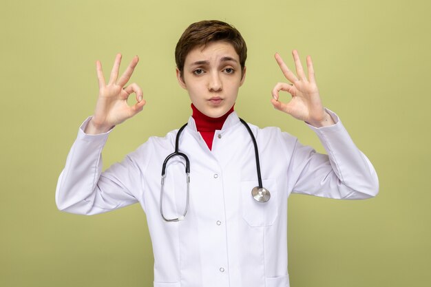 Jeune fille médecin en blouse blanche avec stéthoscope autour du cou souriant confiant faisant signe ok debout sur vert