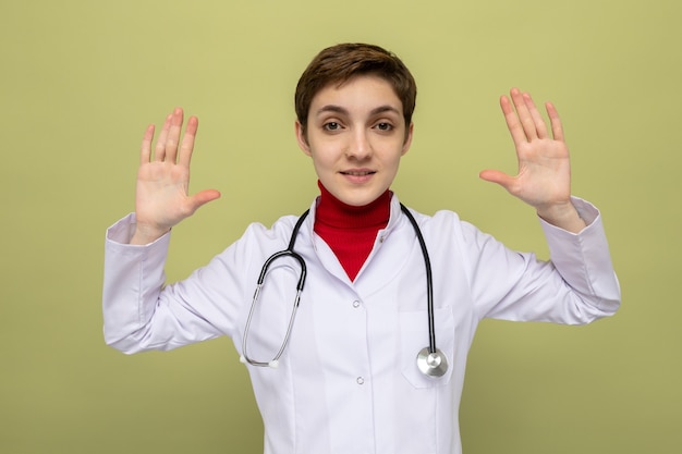 Jeune fille médecin en blouse blanche avec stéthoscope autour du cou souriant confiant levant les bras en se rendant debout sur le vert