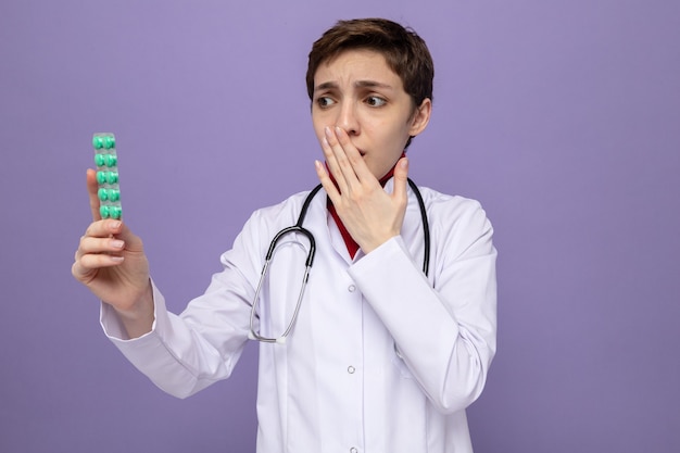 Photo gratuite jeune fille médecin en blouse blanche avec stéthoscope autour du cou tenant une ampoule avec des pilules en le regardant la bouche inquiète avec la main debout sur le violet