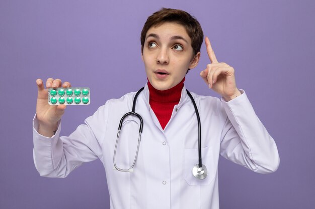 Jeune fille médecin en blouse blanche avec stéthoscope autour du cou tenant un blister avec des pilules jusqu'à surpris montrant l'index debout sur violet