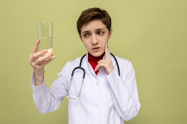 Jeune fille médecin en blouse blanche avec stéthoscope autour du cou tenant un verre d'eau en le regardant perplexe debout sur le vert