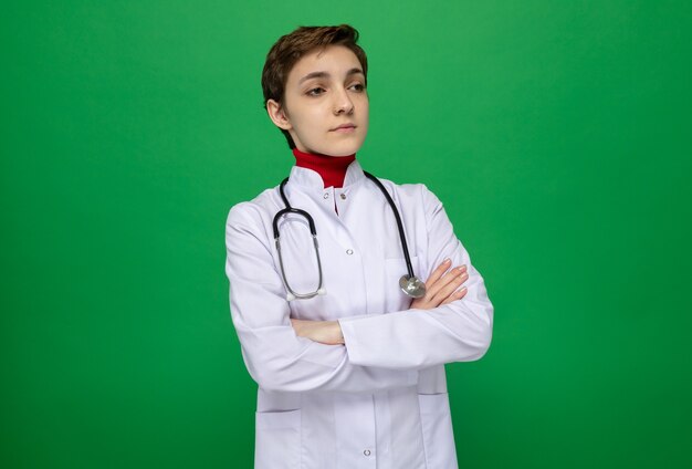 Jeune fille médecin en blouse blanche avec stéthoscope regardant de côté avec une expression sérieuse et confiante avec les bras croisés debout sur le vert
