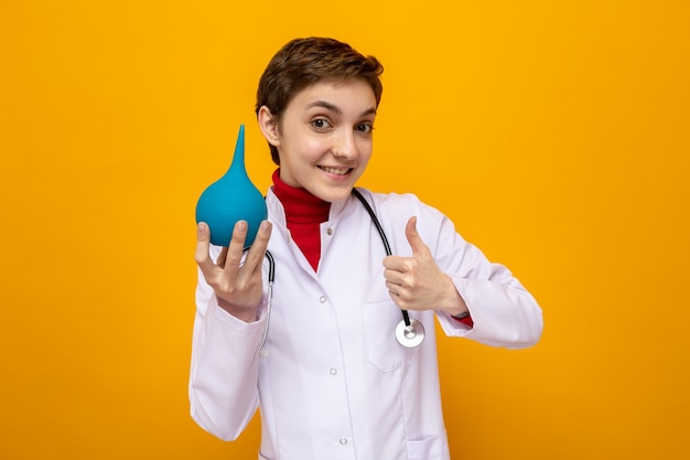 Jeune fille médecin en blouse blanche avec stéthoscope tenant des poires médicales souriant joyeusement montrant les pouces vers le haut debout sur orange