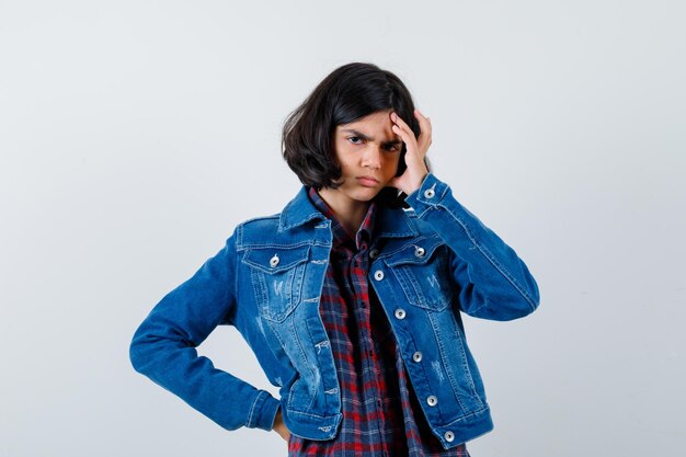 Jeune fille mettant la main sur le front tout en tenant la main sur la taille en chemise à carreaux et veste en jean et regardant pensive, vue de face.