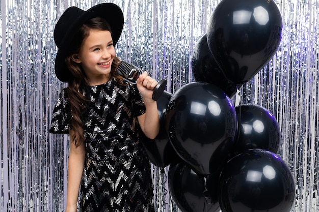 La jeune fille à la mode chante en karaoké lors d'une fête avec un microphone dans ses mains sur un brillant avec des ballons d'hélium noir