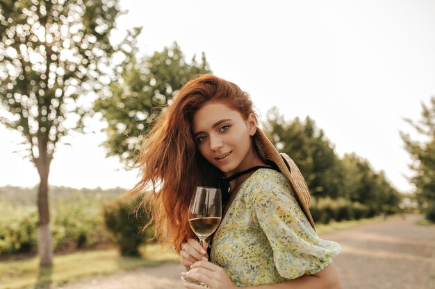 Jeune fille à la mode avec une longue coiffure ondulée au gingembre dans des vêtements d'été imprimés et un chapeau de paille regardant à l'avant et tenant un verre avec du vin en plein air