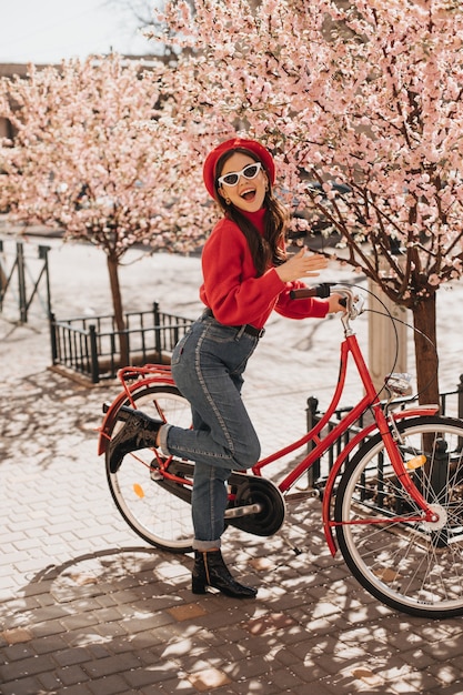 Jeune fille à la mode posant avec vélo rouge près de fleurs de cerisier. Femme en pull en laine et jeans souriant contre sakura