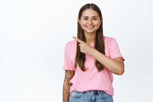 Jeune fille moderne avec bras tatoué, pointant le doigt vers la gauche, montrant la direction sur blanc isolé.