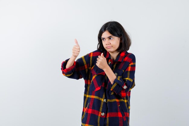 Jeune fille montrant le double pouce vers le haut en chemise à carreaux et l'air heureux, vue de face.