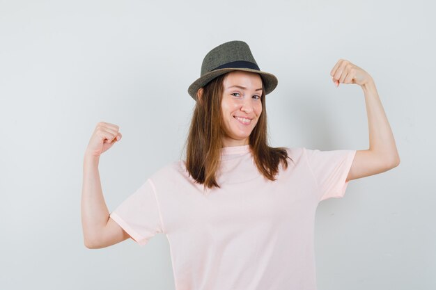 Jeune fille montrant le geste du gagnant en t-shirt rose, chapeau et à la joyeuse. vue de face.