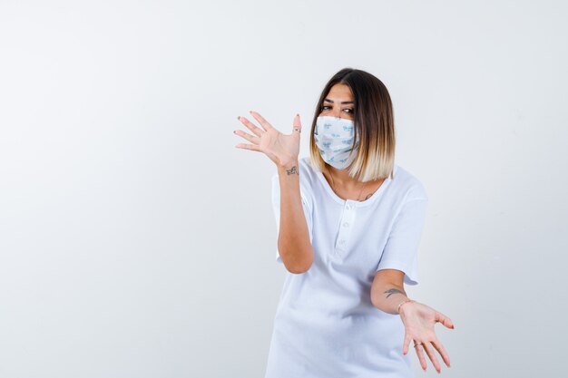 Jeune fille montrant un geste impuissant en t-shirt blanc et masque et à la perplexe, vue de face.