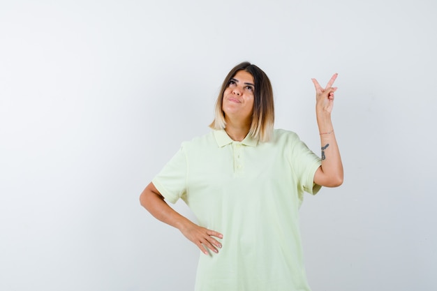 Jeune fille montrant le geste de paix, tenant la main sur la taille en t-shirt et regardant pensif, vue de face.