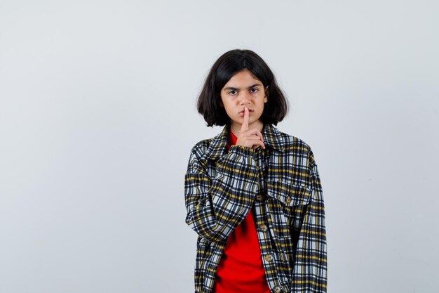 Jeune fille montrant un geste de silence en chemise à carreaux et t-shirt rouge et l'air sérieux, vue de face.