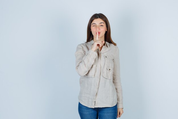 Jeune fille montrant un geste de silence, un clin d'œil en chemise beige, un jean et l'air mignon. vue de face.