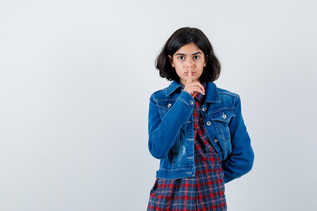 Jeune fille montrant un geste de silence, tenant la main derrière la taille en chemise à carreaux et veste en jean et l'air mignon. vue de face.