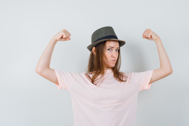 Jeune fille montrant les muscles des bras en t-shirt rose, chapeau et à la confiance. vue de face.