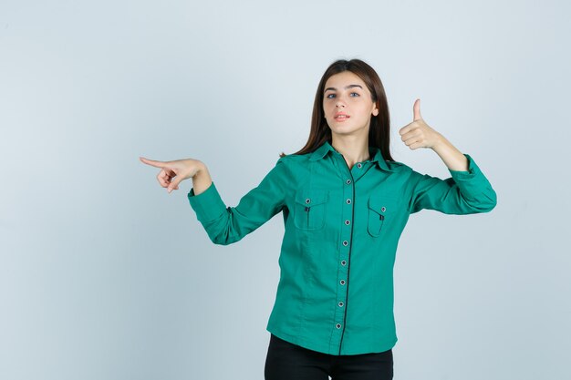 Jeune fille montrant le pouce vers le haut, pointant vers la gauche avec l'index en chemisier vert, pantalon noir et regardant confiant, vue de face.