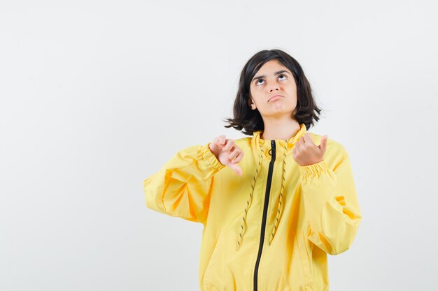 Jeune fille montrant les pouces vers le bas et pensant à quelque chose en blouson aviateur jaune et à la pensif