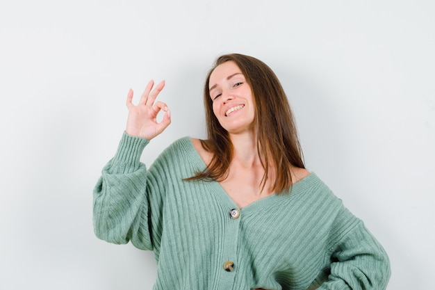 Jeune fille montrant le signe ok, tenant une main sur la taille en maille et à la confusion, vue de face.