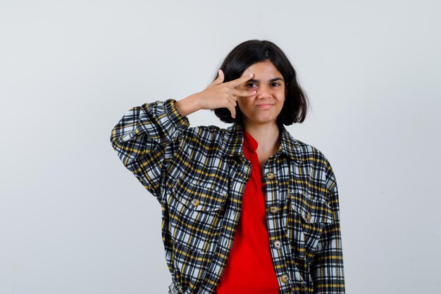 Jeune fille montrant un signe v sur l'œil en chemise à carreaux et t-shirt rouge et l'air heureux. vue de face.