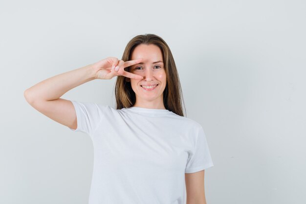 Jeune fille montrant le signe v sur l'oeil en t-shirt blanc et à la confiance. vue de face.