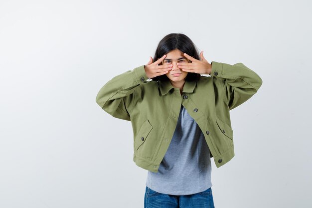 Jeune fille montrant des signes v sur les yeux en pull gris, veste kaki, pantalon en jean et l'air sérieux. vue de face.