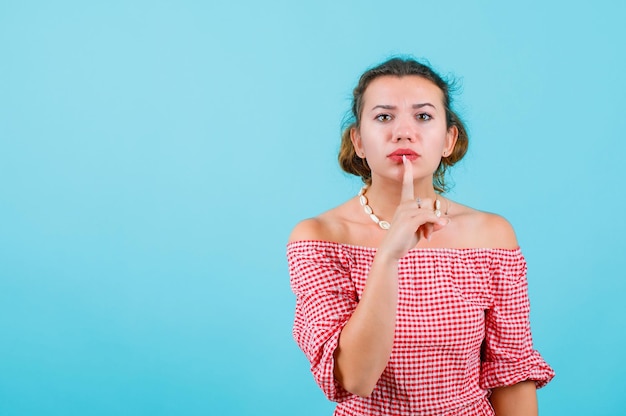 Jeune fille montre un geste de silence en tenant l'index sur les lèvres sur fond bleu