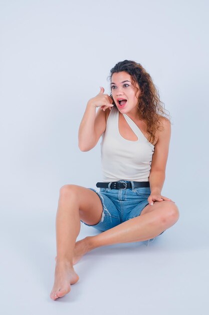 Jeune fille montre un geste de téléphone avec la main en s'asseyant sur le sol sur fond blanc