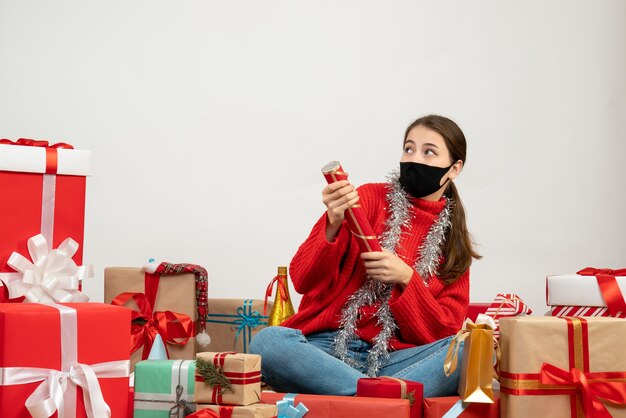 Jeune fille de Noël avec masque noir tenant popper party assis autour de cadeaux sur blanc
