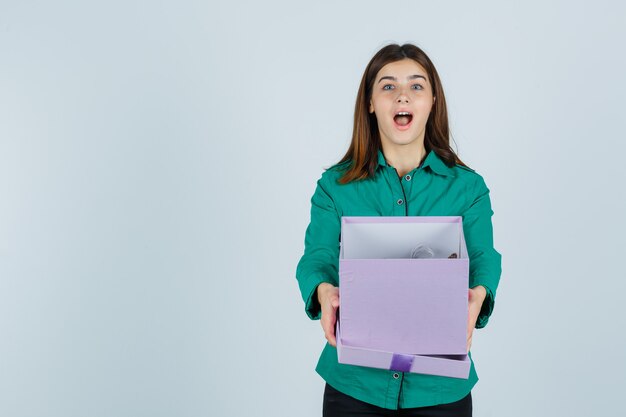 Jeune fille ouvrant la boîte-cadeau, gardant la bouche grande ouverte en chemisier vert, pantalon noir et à la surprise. vue de face.