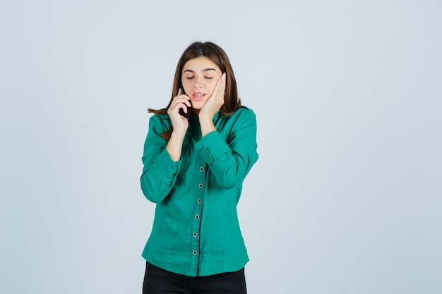 Jeune fille parlant au téléphone, tenant la main sur la joue en chemisier vert, pantalon noir et regardant curieux, vue de face.