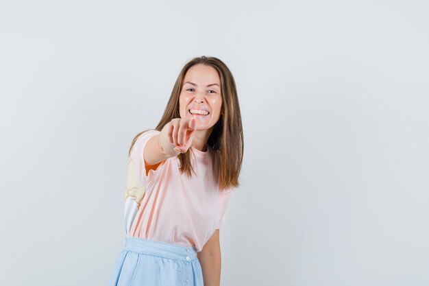 Jeune fille pointant la caméra en t-shirt, jupe et à la joyeuse. vue de face.